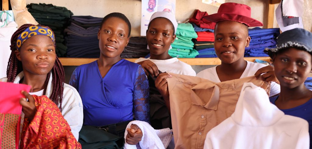 Grupp av leende unga kvinnor som visar upp nyss färdigställda klädesplagg i en textilbutik, med hyllor av tyg i bakgrunden.
